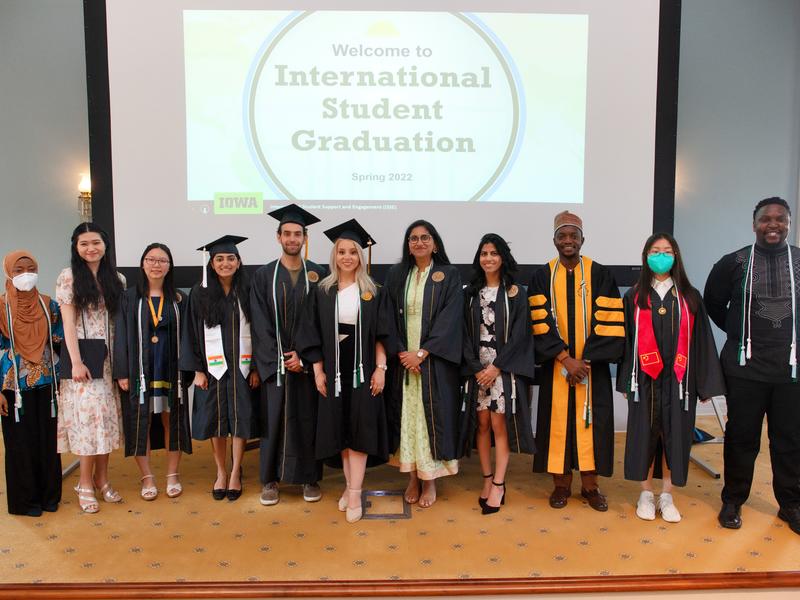a group of international students wearing the international student graduation cord standing in front of a screen says "welcome to international student graduation" 